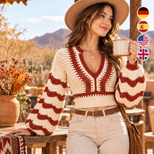 May include: A long-sleeved crochet sweater featuring a cream and rust-colored wavy pattern, a deep V-neckline, and a fitted waist. The sweater is paired with light-colored denim jeans and a brown leather belt with a gold-toned buckle.