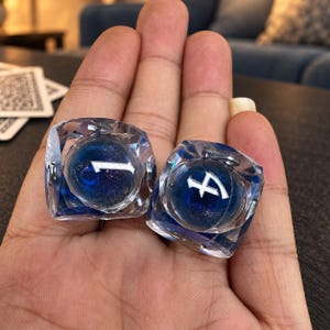 May include: Two transparent, multi-faceted dice with a deep blue, swirling core and glittery accents. Each die features a large, white, stylized numeral on its face, with one displaying the number one and the other displaying the number four.