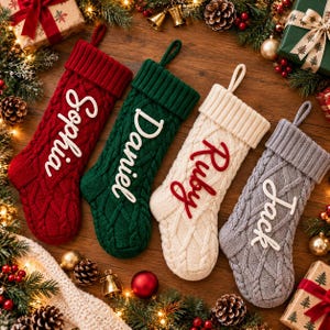 May include: Four cable-knit Christmas stockings with hanging loops, each featuring a name in raised script lettering. The stockings are red with white text, green with white text, cream with red text, and gray with white text.