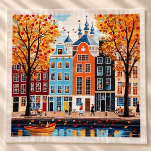 May include: A cross-stitch embroidery depicting a canal-side city scene with colorful buildings in red, blue, orange, and black. Two large trees with orange autumn leaves frame the scene, while a small boat floats on the water below.