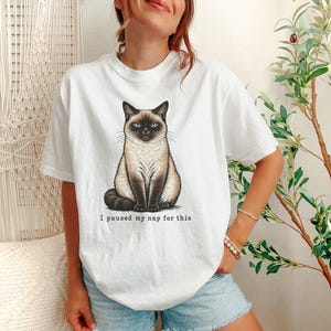 May include: A white short-sleeved t-shirt featuring a graphic illustration of a grumpy Siamese cat sitting upright. Below the cat, the text reads, I paused my nap for this. The design is centered on the chest of the garment.