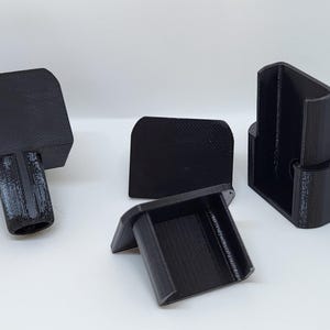 Op de afbeelding: Een set van vier zwarte 3D-geprinte kunststof onderdelen, elk voorzien van een platte rechthoekige basis bevestigd aan een holle, U-vormige beugel of een cilindrische paal, met een getextureerde matte afwerking en stijve constructie.