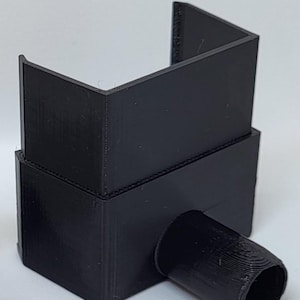 May include: A black 3D-printed plastic component featuring a rectangular base with a protruding cylindrical port on the front side and an open-topped U-shaped vertical extension rising from the top surface, showing visible layer lines.