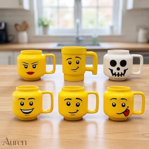 Puede incluir: Una colección de seis tazas de cerámica con forma de cabezas de juguete. Cinco tazas son de color amarillo brillante con varias expresiones faciales impresas, incluyendo caras sonrientes y neutras. Una taza es blanca con un diseño de calavera.