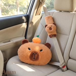 Peut inclure: Un coussin en peluche en forme de capybara beige avec un petit fruit orange sur le dessus, doté d'oreilles marron foncé et d'un visage brodé. Une housse de ceinture de sécurité assortie en peluche capybara est fixée sur la sangle beige.