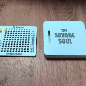 May include: A white XP Tracker board with a grid of holes, numbers 0-9, increments of +10 to +90, and three yellow pegs, alongside a white square case featuring the text THE SAVAGE SOUL and the website savagesoul.com printed on the tracker.