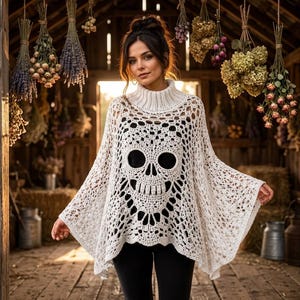 Puede incluir: Un poncho de ganchillo blanco con un motivo de calavera prominente en la parte delantera. La prenda tiene un cuello alto de canalé y un intrincado patrón de encaje calado en todo el cuerpo, creando una silueta holgada y fluida sobre ropa negra.