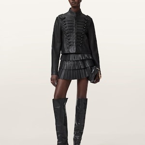 May include: A black leather military-style jacket with braided cord detailing and buttons, paired with a black tiered pleated mini skirt and black leather knee-high boots. A small black clutch with a gold chain strap is held in one hand.