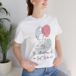 May include: A white short-sleeve t-shirt featuring a black line art illustration of a person sitting with long wavy hair. Behind the figure are abstract pink and grey shapes. The text reads, "Be still and know That I am God." Psalm 46:10.