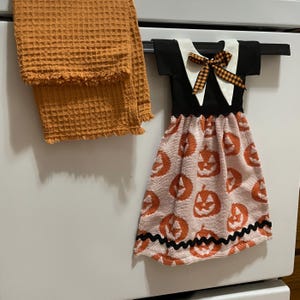 以下が含まれることがあります： オレンジ色のワッフル織りのキッチンタオルと、装飾用の吊り下げタオル。吊り下げタオルはドレスのようにデザインされており、黒いトップス、白い襟、カボチャ模様のスカートが付いています。黒とオレンジのリボンが上部を飾っています。