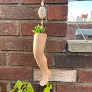 May include: A unique hanging planter shaped like a doll's leg, holding a small succulent. The planter is suspended by a gold chain with decorative beads. The background is a brick wall and a cactus.