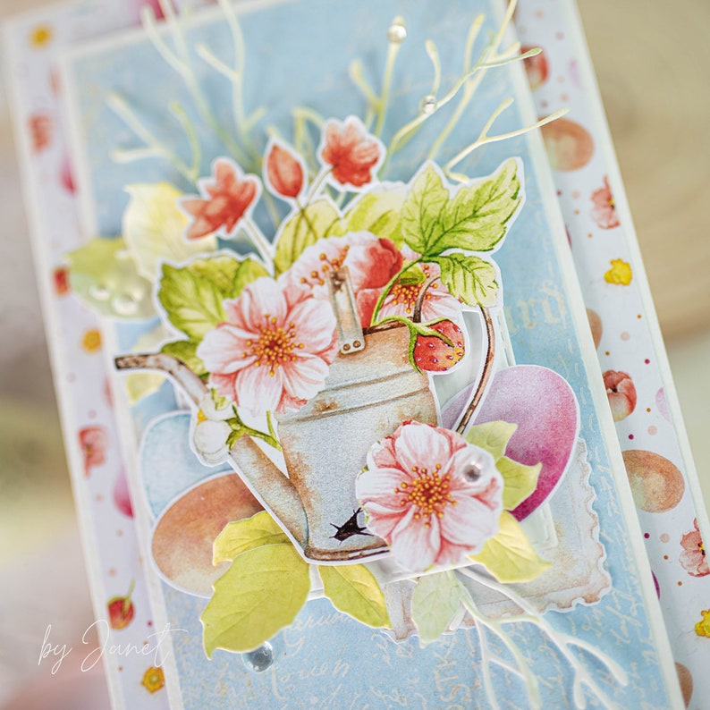 May include: A blue and white card with a watercolor illustration of a watering can with pink flowers and green leaves. The card has a white border and the text "by Janet" in the bottom left corner.