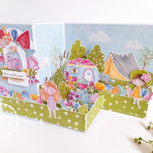 May include: A pop-up card with a camping theme. The card features a camper, a tent, trees, and flowers. The card is decorated with a blue sky and white clouds. The text on the card reads "Nasz wsp&oacute;lny czas".