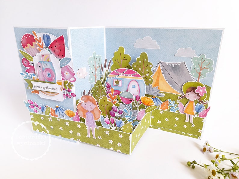 May include: A pop-up card with a camping theme. The card features a camper, a tent, trees, and flowers. The card is decorated with a blue sky and white clouds. The text on the card reads "Nasz wsp&oacute;lny czas".