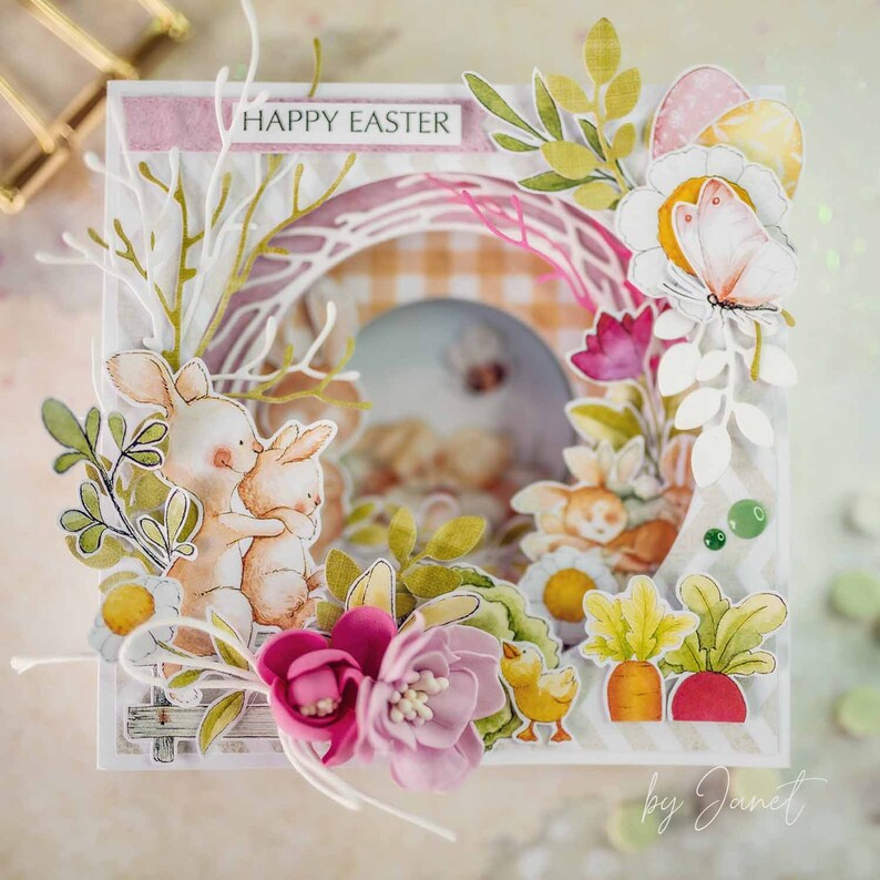 May include: A handmade Easter card with a pop-up design featuring two bunnies hugging, surrounded by flowers, greenery, and a checkered background. The card is decorated with a white lace frame and the words "Happy Easter" in pink lettering.