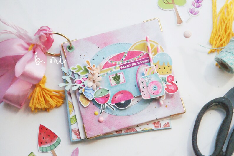 May include: A pink and blue scrapbook with a camper van, a flamingo, and a watermelon on the cover. The text "Wakacyjne Wyjazdy" is on the camper van.