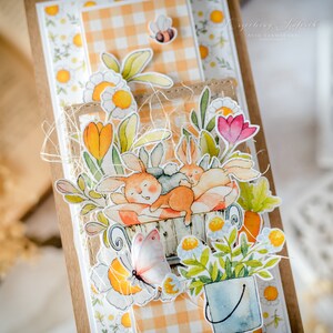 May include: A handmade card with a watercolor illustration of two bunnies sleeping in a basket surrounded by flowers and a butterfly. The card has a gingham pattern and the text "Greeting Kalendar" is visible.