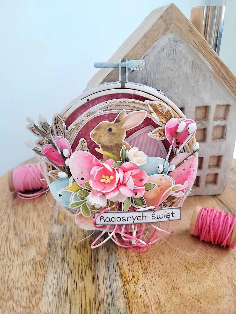 May include: A handmade Easter craft featuring a brown bunny rabbit surrounded by pastel pink, blue, and yellow eggs. The craft is decorated with flowers and greenery and includes the text "Radosnych Świąt" which translates to "Happy Holidays" in English.