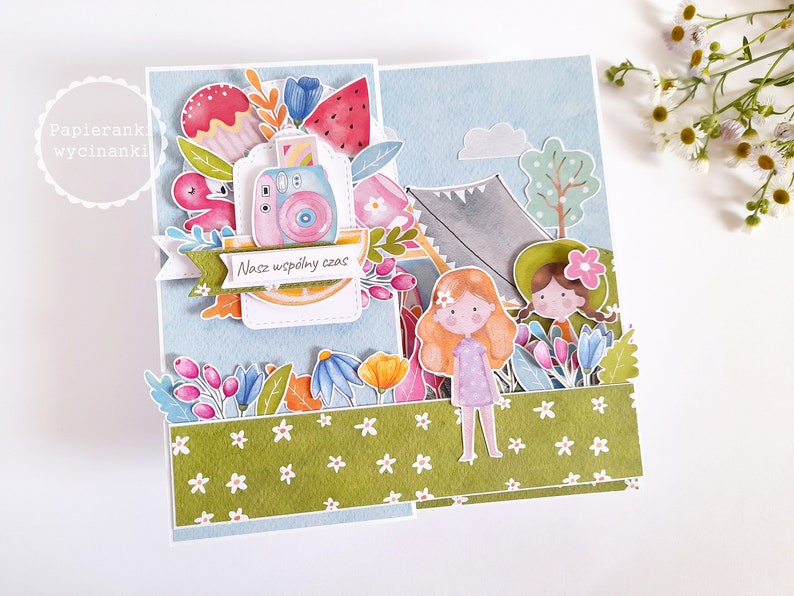 May include: A colorful card with a blue background and a green border. The card features a watercolor illustration of two girls camping with a tent, a camera, and flowers. The text on the card reads "Nasz wsp&oacute;lny czas".