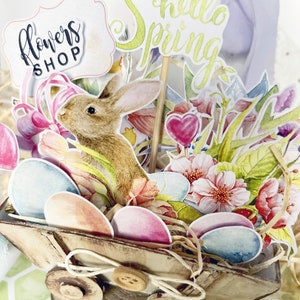May include: A wooden wagon filled with pastel colored eggs, flowers, and a bunny rabbit. The wagon is decorated with a sign that says "Flowers Shop" and a sign that says "Hello Spring".