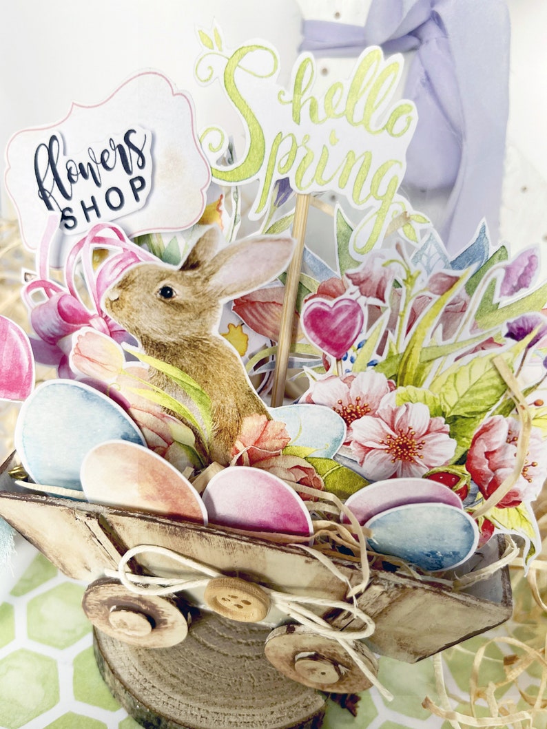 May include: A wooden wagon filled with pastel colored eggs, flowers, and a bunny rabbit. The wagon is decorated with a sign that says "Flowers Shop" and a sign that says "Hello Spring".