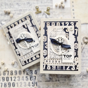 Peut inclure: Deux petits carnets recouverts de tissu blanc avec des chiffres noirs et un motif de clé. Les carnets ont un ruban noir et une étiquette qui dit "Top Secret".