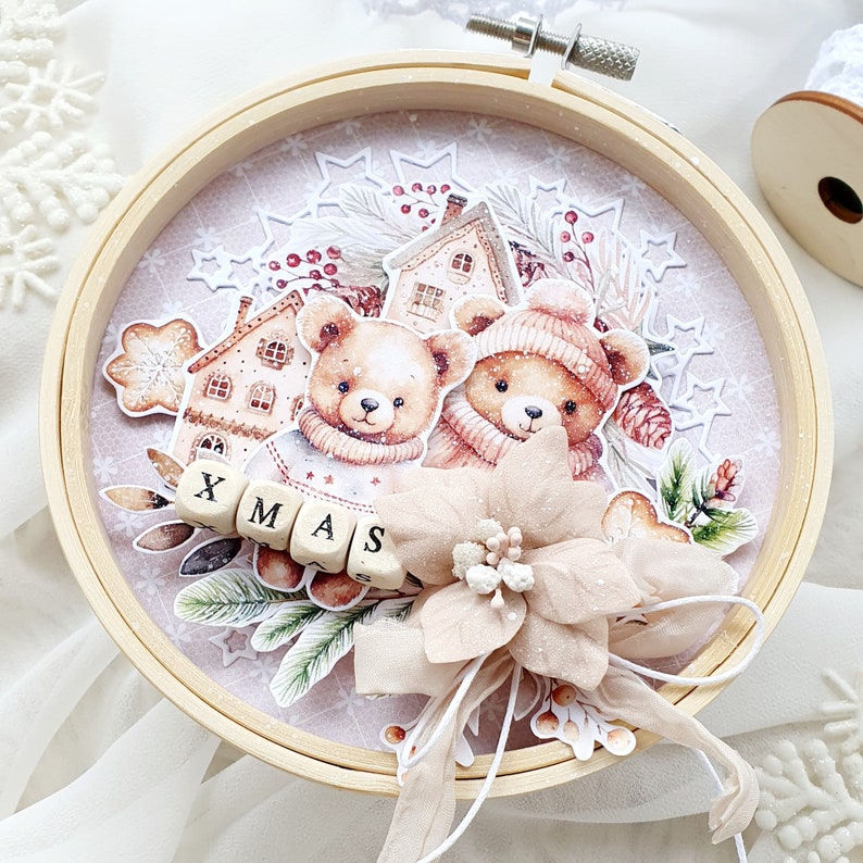 May include: A wooden round frame with a pink background featuring two brown teddy bears wearing winter hats and scarves. The bears are surrounded by a winter scene with a small house, snowflakes, and a poinsettia flower. The frame has the word "XMAS" spelled out in wooden blocks.