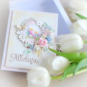 May include: A white card with a pastel floral design and the word "Alleluya" written in white script. The card is surrounded by white tulips.