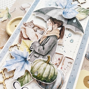 Op de afbeelding: Een aquarel-illustratie van een vrouw die een paraplu vasthoudt, een kat die in een mand slaapt en een pompoen. De illustratie is op een witte achtergrond met blauwe en bruine accenten.