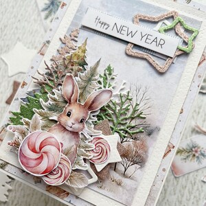 May include: A handmade greeting card with a winter scene featuring a brown bunny, a green pine tree, and a white banner with the text "Happy New Year".