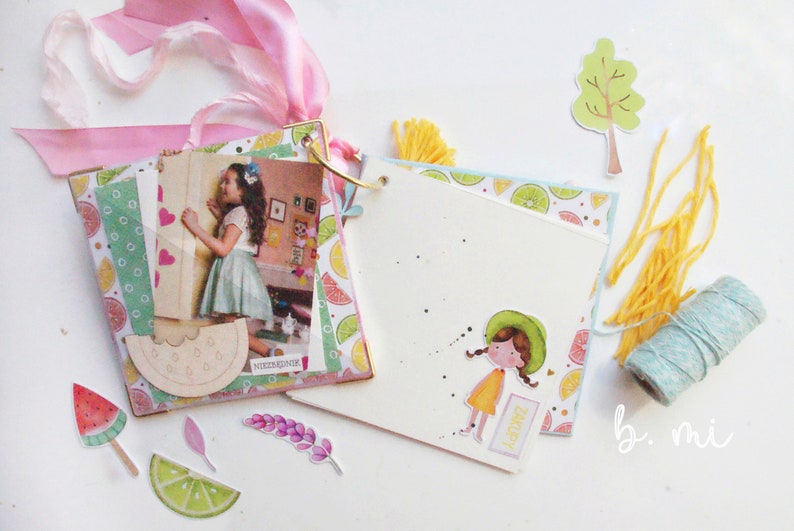 May include: A scrapbook with a colorful lemon and lime pattern, a photo of a girl, and a watermelon slice cutout. The scrapbook is decorated with pink ribbon, yellow yarn, and a small tree cutout. The scrapbook is open to a blank page with a sticker of a girl in a yellow dress and a green hat. The page also has the word "Happy" written on it.