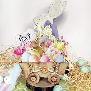 May include: A wooden wagon filled with pastel colored Easter eggs, a brown bunny, and flowers. The wagon has a sign that says "Flowers Shop" and a sign that says "Hello Spring".