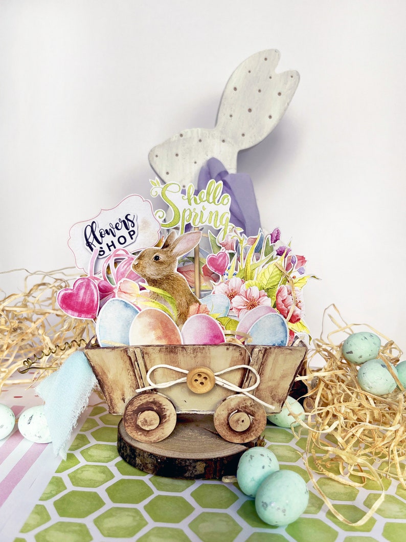 May include: A wooden wagon filled with pastel colored Easter eggs, a brown bunny, and flowers. The wagon has a sign that says "Flowers Shop" and a sign that says "Hello Spring".