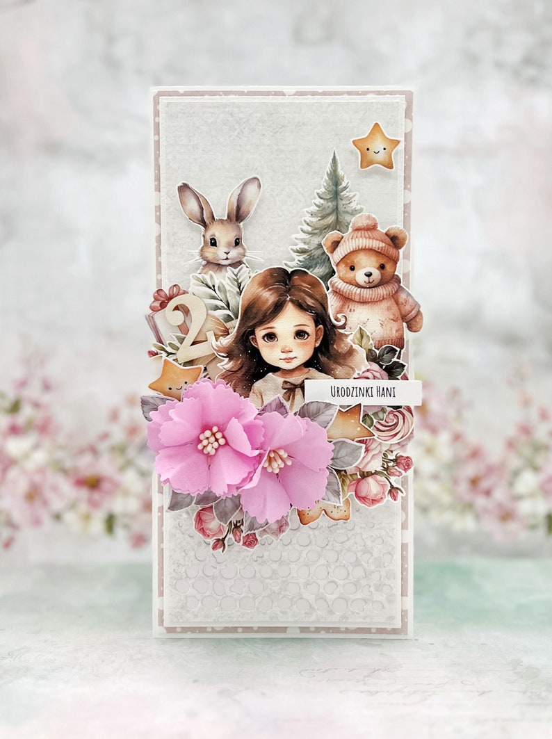 May include: A white card with a pink and white floral design. The card features a girl with brown hair, a bunny, a teddy bear, and a Christmas tree. The text "Urodzinki Hani" is written on the card.