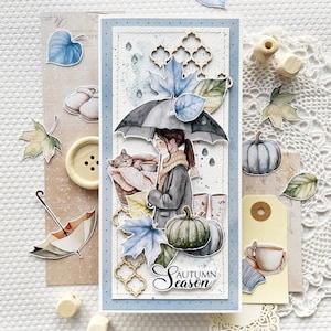 Op de afbeelding: Een handgemaakte kaart met een aquarel-illustratie van een vrouw met een kat onder een paraplu. De kaart heeft een blauwe en witte achtergrond met een bruine en gouden gepatenteerde rand. De tekst "Herfstseizoen" staat in cursief op de kaart.