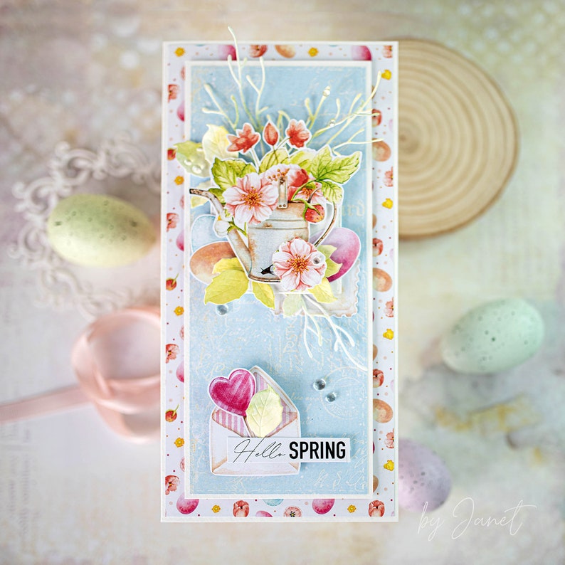 May include: A handmade card with a blue background featuring a watercolor illustration of a watering can with flowers and leaves. The card has a white envelope with a pink heart and the text "Hello Spring" on it.