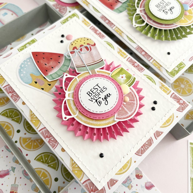 May include: A white card with a pink and white rosette with the text "Best Wishes to you" in black. The card is decorated with images of watermelon, ice cream, and lemons. The card is sitting on a box with a lemon and lime pattern.