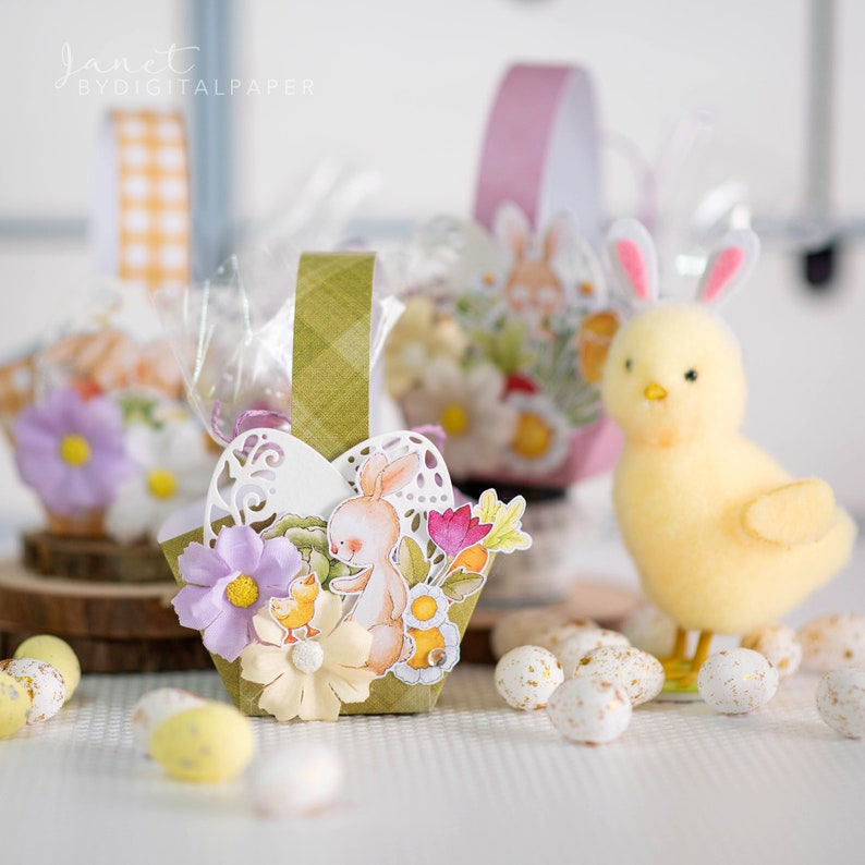May include: A green and white checkered paper basket with a bunny and flowers. The basket is filled with small white and gold speckled eggs. The basket is sitting on a white surface with a yellow chick.