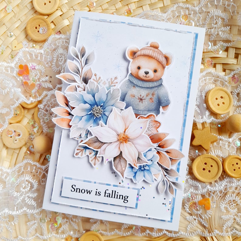 May include: A white card with a watercolor illustration of a brown bear wearing a blue sweater and a white hat. The bear is surrounded by blue and white flowers. The card has the text "Snow is falling" on it.