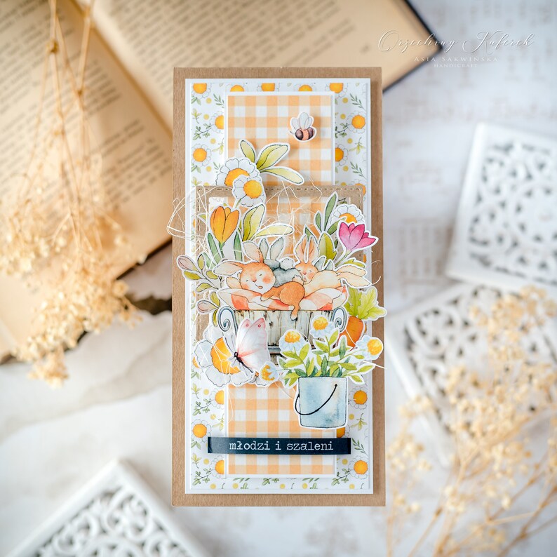 May include: A handmade card with a brown background and a white and orange checkered pattern. The card features a watercolor illustration of two bunnies in a bucket with flowers and a butterfly. The text "młodzi i szaleni" is written on the card.
