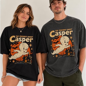 May include: A black short-sleeve t-shirt featuring a graphic print of the cartoon character Casper the Friendly Ghost flying above a haunted house. The design includes a large orange moon, bats, and stars, with the text The Friendly Ghost Casper.