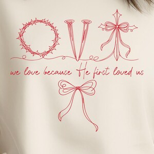 May include: Cream-colored sweatshirt with a red line drawing of a crown of thorns, nails, a cross, and bows. The text "we love because He first loved us" is below the drawings. The bottom of the image says "DIGITAL DOWNLOAD".