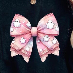 Puede incluir: Una camiseta negra con un lazo rosa y blanco con ribete de encaje y un patrón de fantasmas blancos. El lazo está centrado en la camiseta.