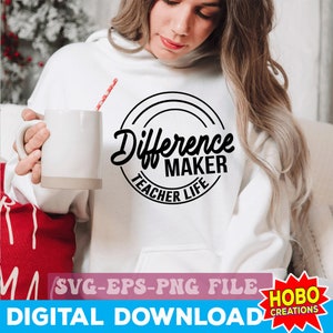 Op de afbeelding: Wit sweatshirt met zwarte tekst die in een cirkel "Difference Maker Teacher Life" luidt. De tekst is in een handschriftlijk lettertype. Het sweatshirt wordt gedragen door een persoon die een witte mok vasthoudt.