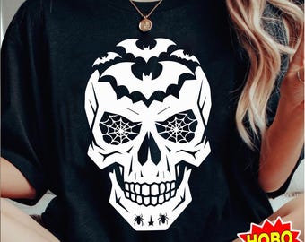 Puede incluir: Camiseta negra con un diseño de calavera blanca. La calavera tiene telarañas por ojos y murciélagos alrededor de la cabeza. También hay pequeñas arañas en la parte inferior del diseño.