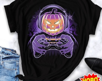 Könnte beinhalten: Ein schwarzes T-Shirt mit einem lila und orangefarbenen Kürbis, der Kopfhörer trägt und einen Videospiel-Controller hält. Der Kürbis hat einen gruseligen Ausdruck und ist vor einem dunklen Hintergrund mit einem Spinnennetz und einem Spukhaus platziert.