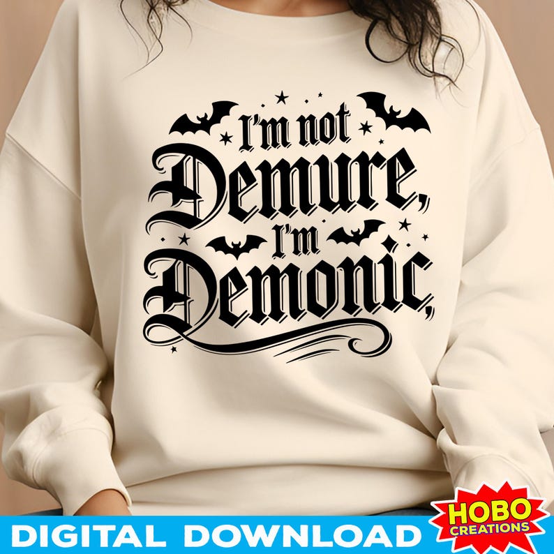 May include: A cream-coloured sweatshirt with a black graphic print that reads "I'm not Demure, I'm Demonic" with bats and stars surrounding the text.