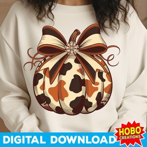 May include: A white sweatshirt featuring a brown and white cow print pumpkin with a brown bow and a rhinestone in the centre. The text "HOBO CREATIONS" is on a red starburst.