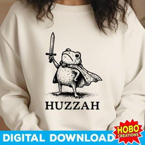 May include: Cream-colored sweatshirt featuring a black and white illustration of a frog holding a sword and wearing a cape. The word "Huzzah" is printed below the frog. The bottom of the image has the text "DIGITAL DOWNLOAD".