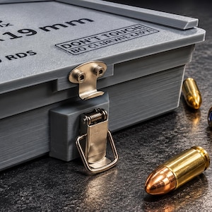 Könnte beinhalten: Eine graue, rechteckige Aufbewahrungsbox mit Metallverschluss, beschriftet mit ARMORIX, 9 x 19 mm, 100 RDS und DON'T TOUCH! BO CI URWIE ŁAPY!. Mehrere messingfarbene Patronen mit Kupferspitzen liegen neben der Box auf einer dunklen Oberfläche.